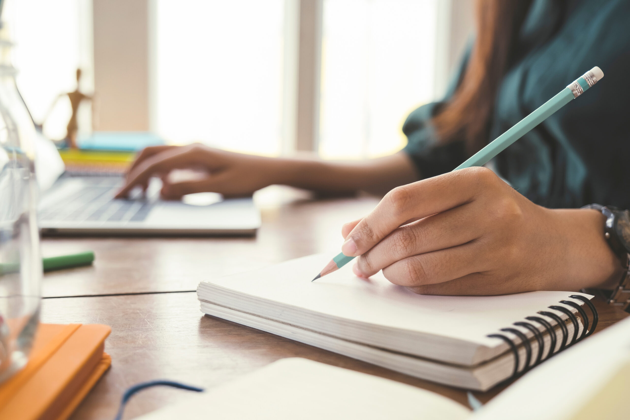 A woman writing with one hand in a notebook with a pencil and with another hand on the computer keyboard.