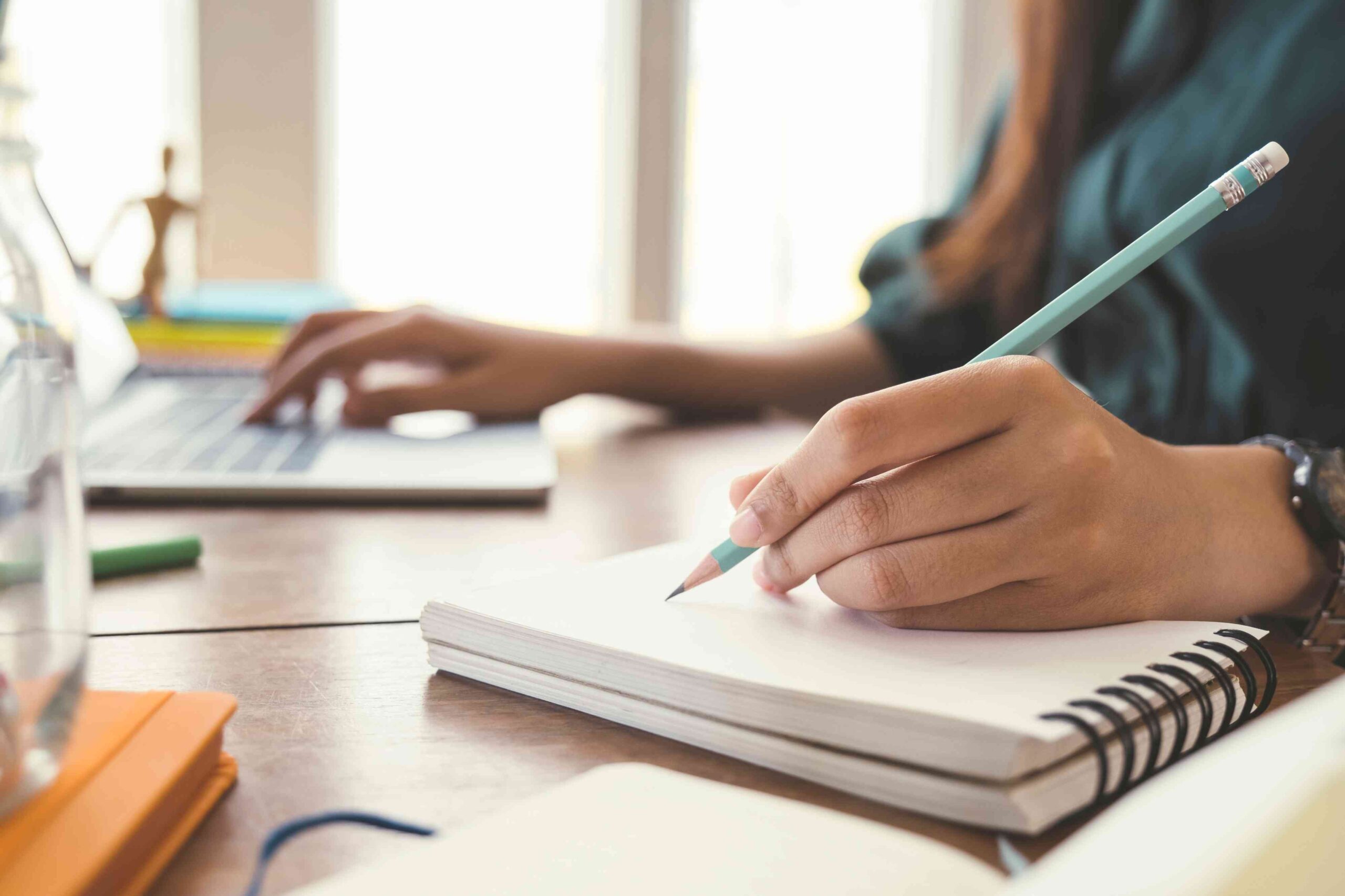 A woman writing with one hand in a notebook with a pencil and with another hand on the computer keyboard.